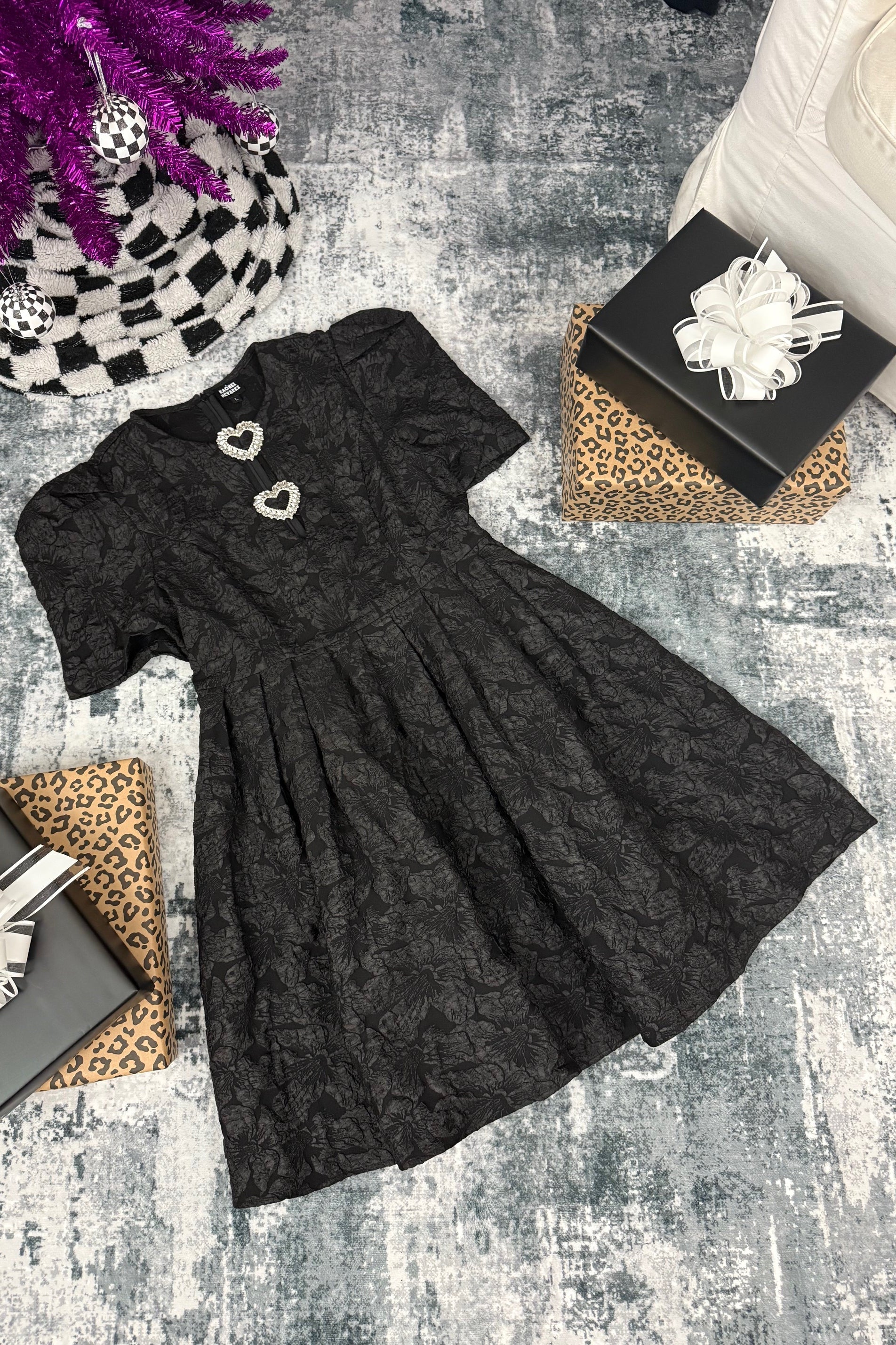 Black dress with decorative buttons on a textured surface with gift boxes and a plant.