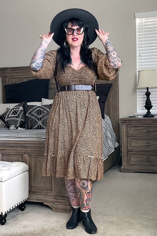A person wearing a brown leather fashion harness with an adjustable waist belt and shoulder straps, featuring metal buckles and ring. The harness is styled with a leopard print dress, black hat, and sunglasses.