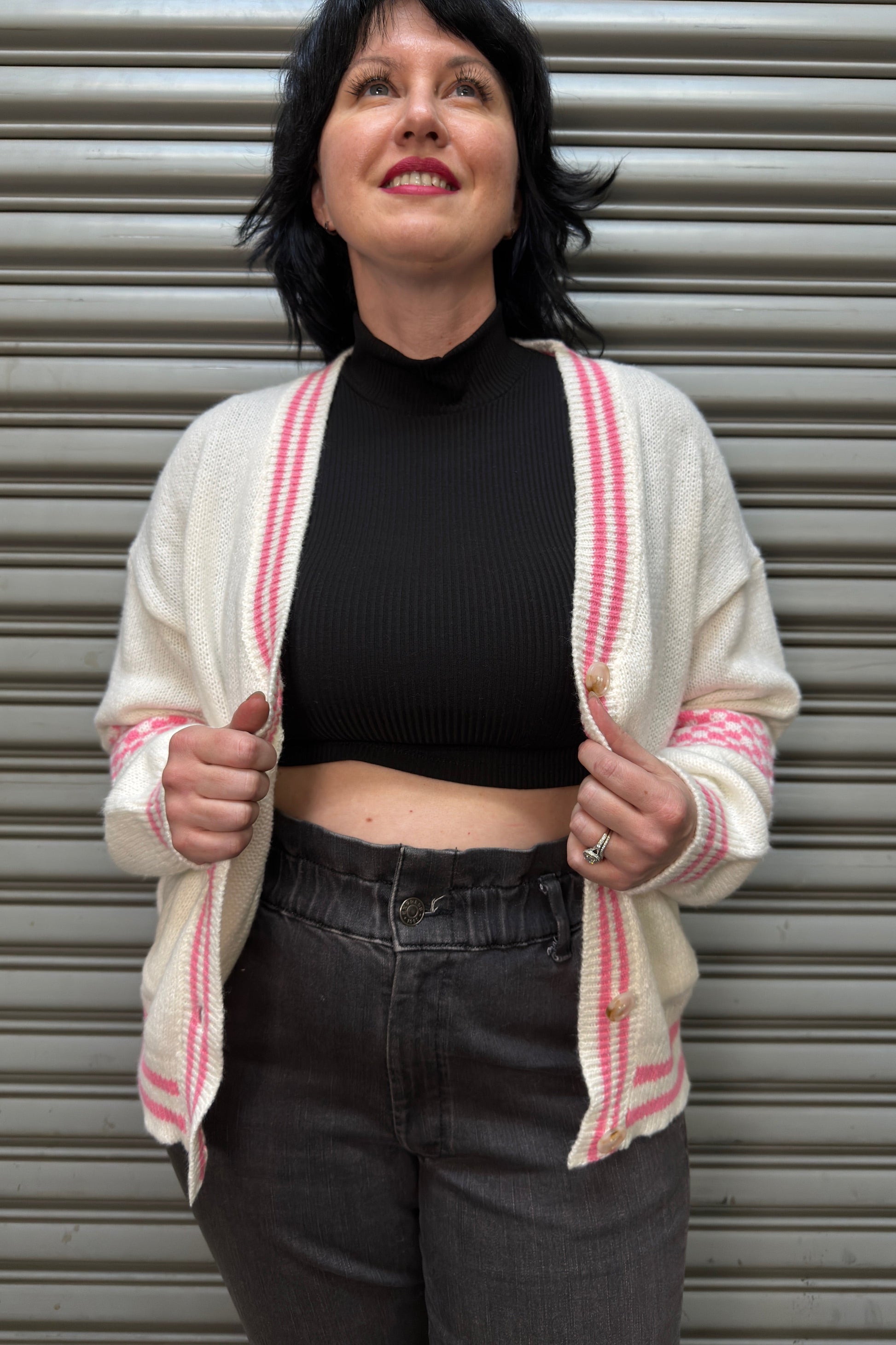Woman wearing a white cardigan with red stripes, black top, and dark jeans in front of a metal shutter.