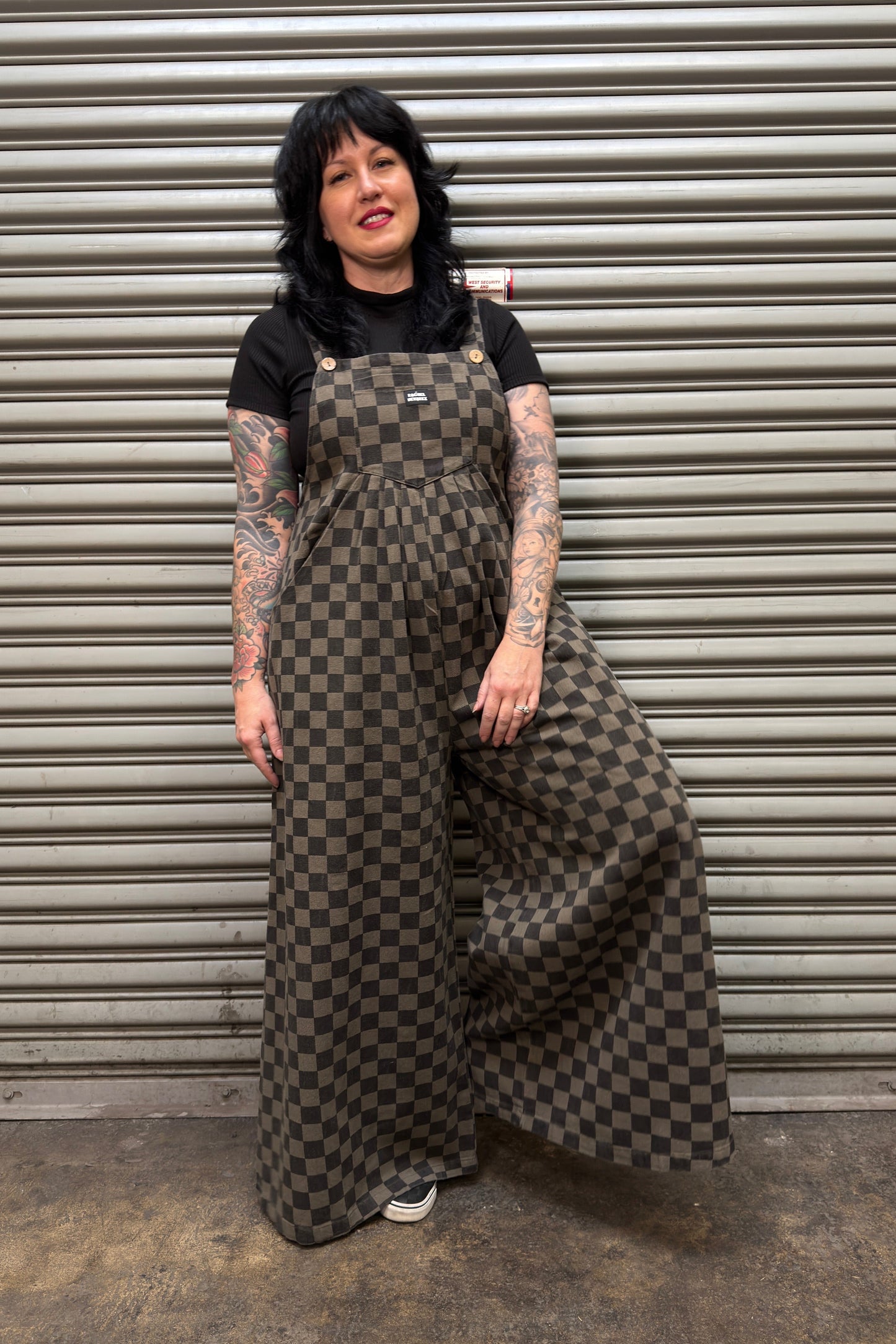 Person wearing a checkered overalls standing in front of a metal shutter.