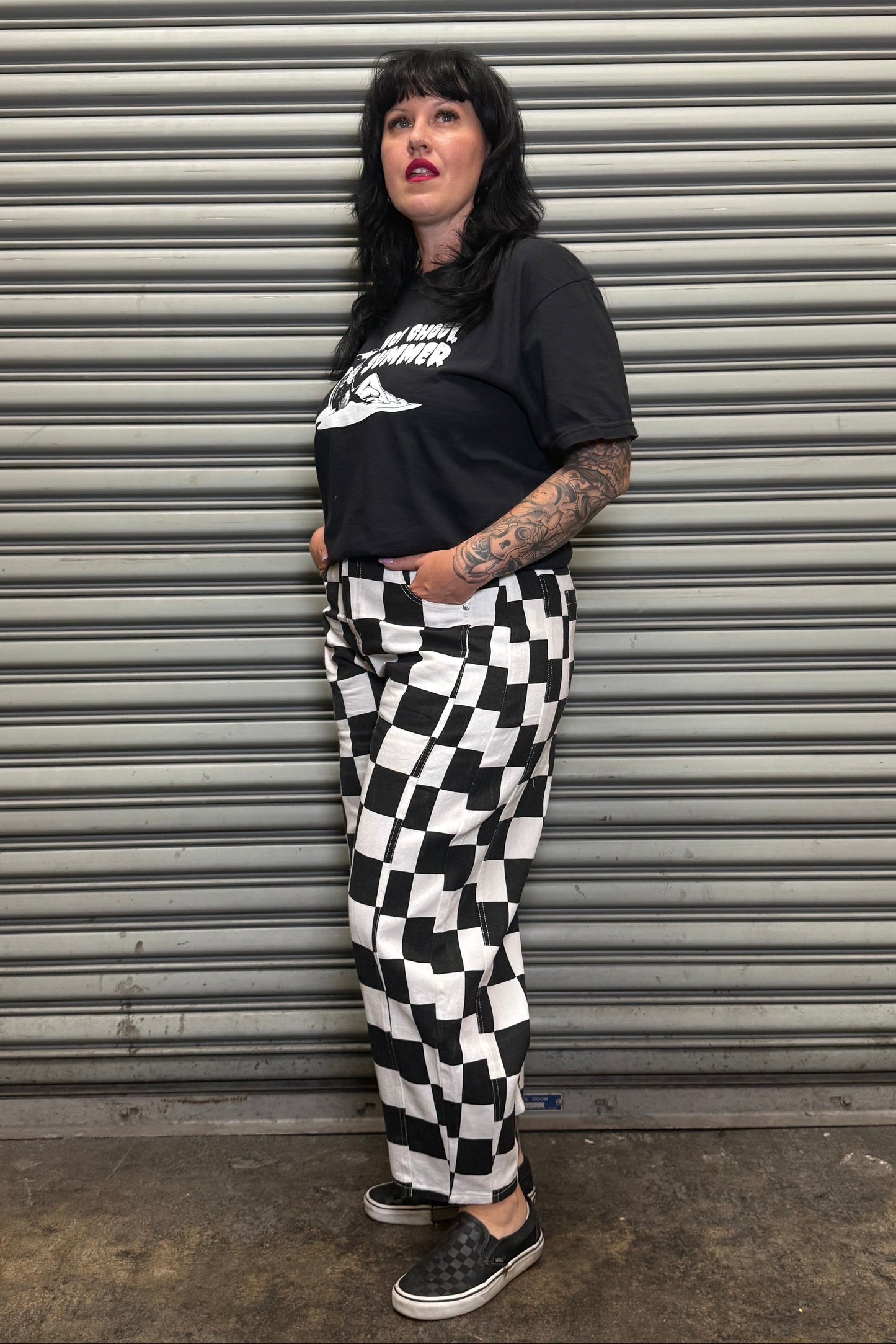 Person wearing a black t-shirt with white text and checkered pants standing against a metal shutter.