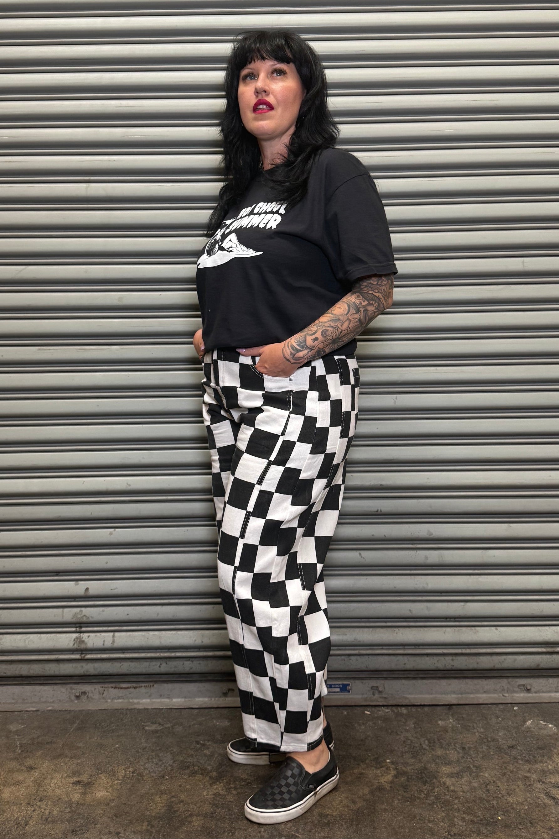 Person wearing a black t-shirt with white text and checkered pants standing against a metal shutter.