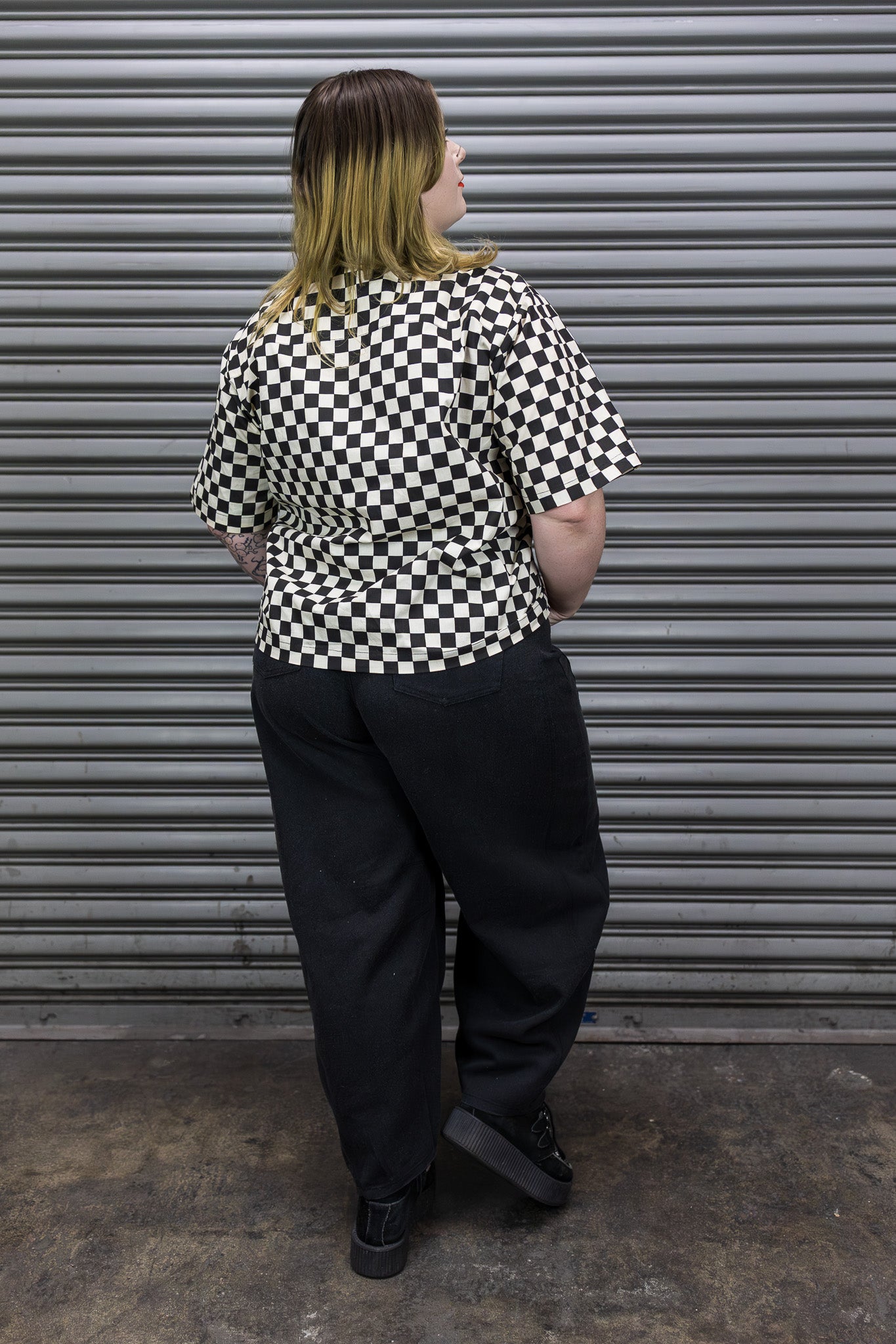 Person wearing a black and white checkered shirt and black pants standing in front of a gray metal shutter.