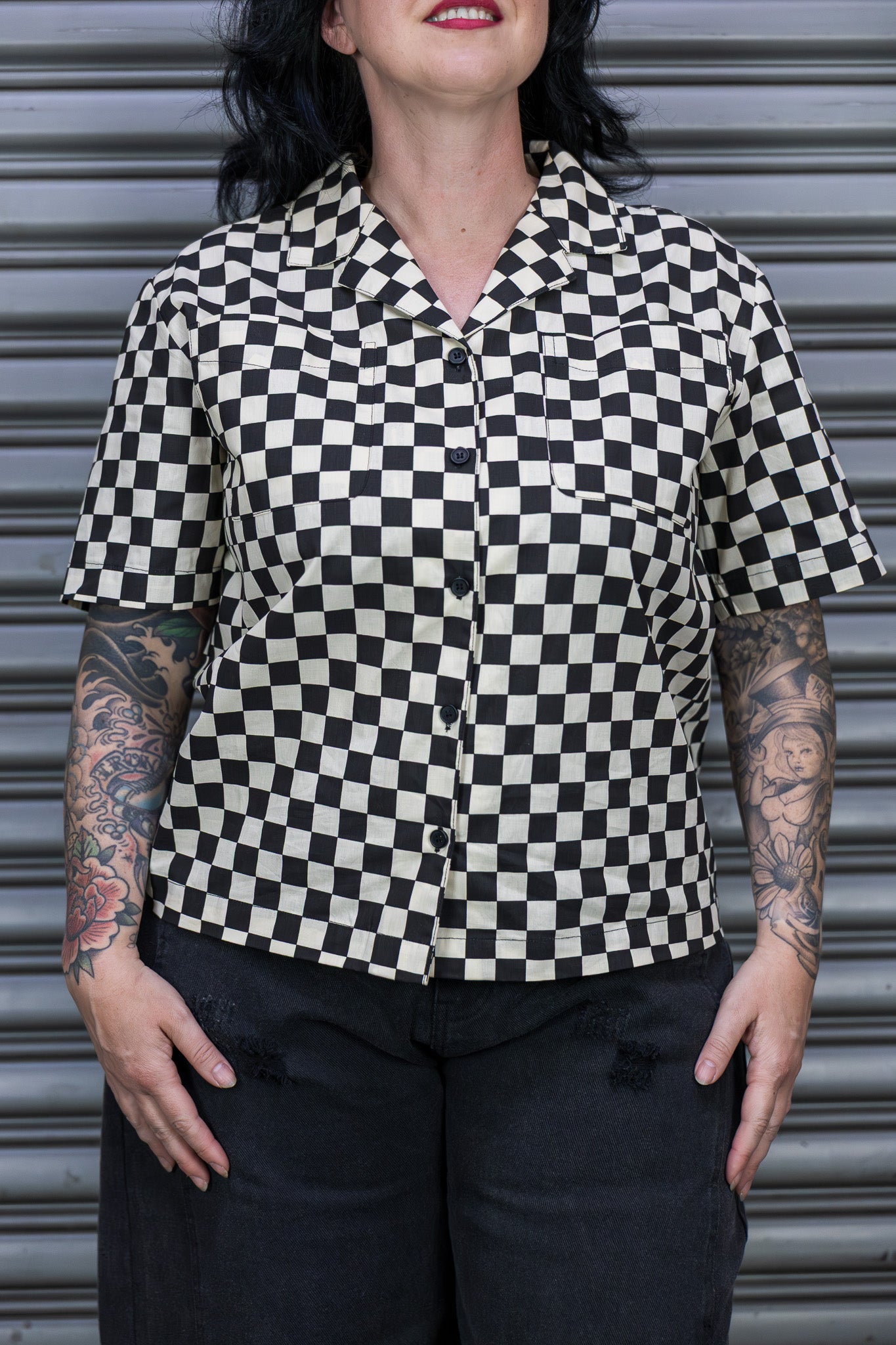 Person wearing a black and white checkered shirt with tattoos visible on arms, standing against a metallic shutter.