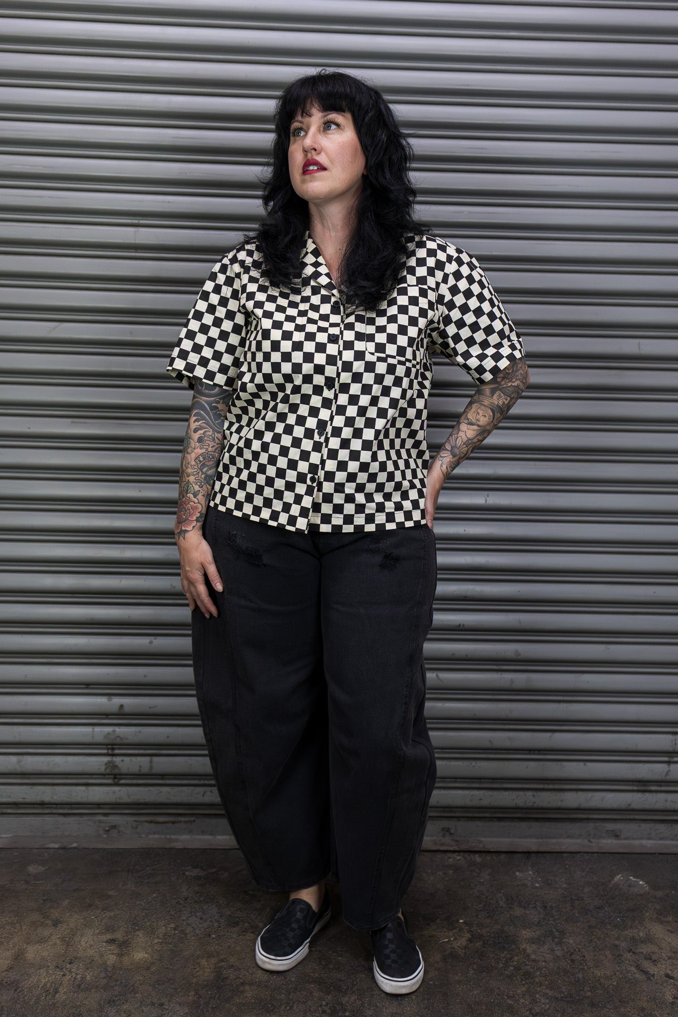 Person wearing a black and white checkered shirt standing in front of a metal shutter.