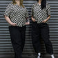 Two women wearing matching black and white checkered shirts and black pants standing against a metallic shutter.