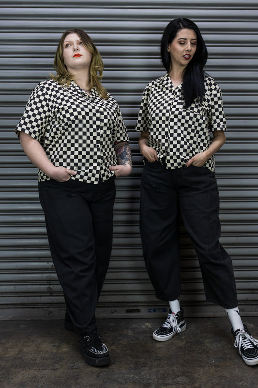 Two women wearing matching black and white checkered shirts and black pants standing against a metallic shutter.