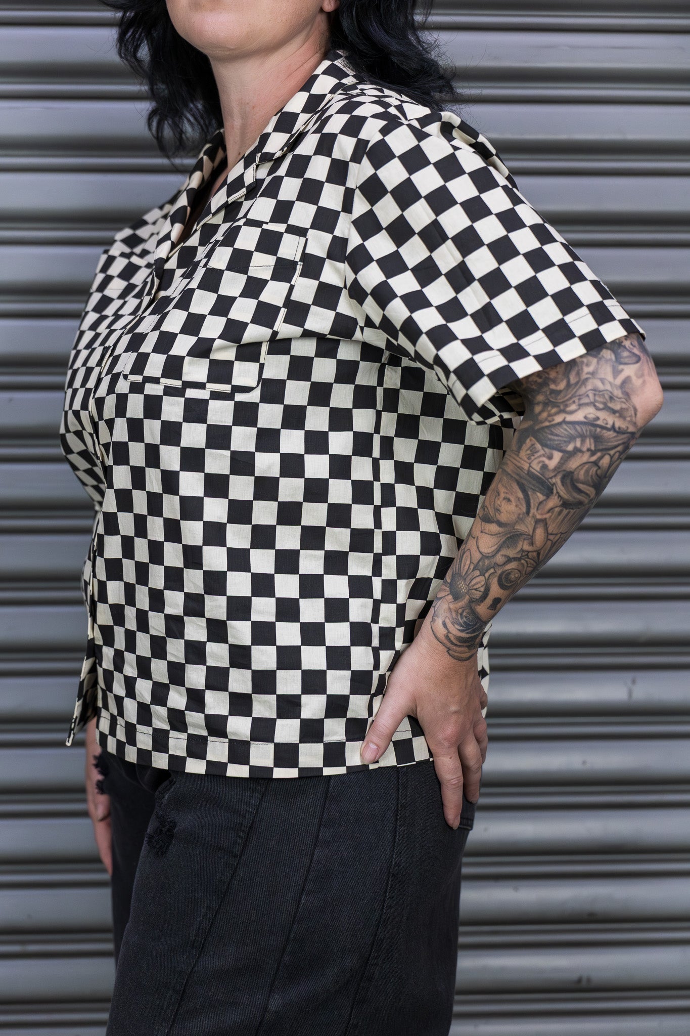 Person wearing a black and white checkered shirt with tattoos, standing in front of a metallic shutter.