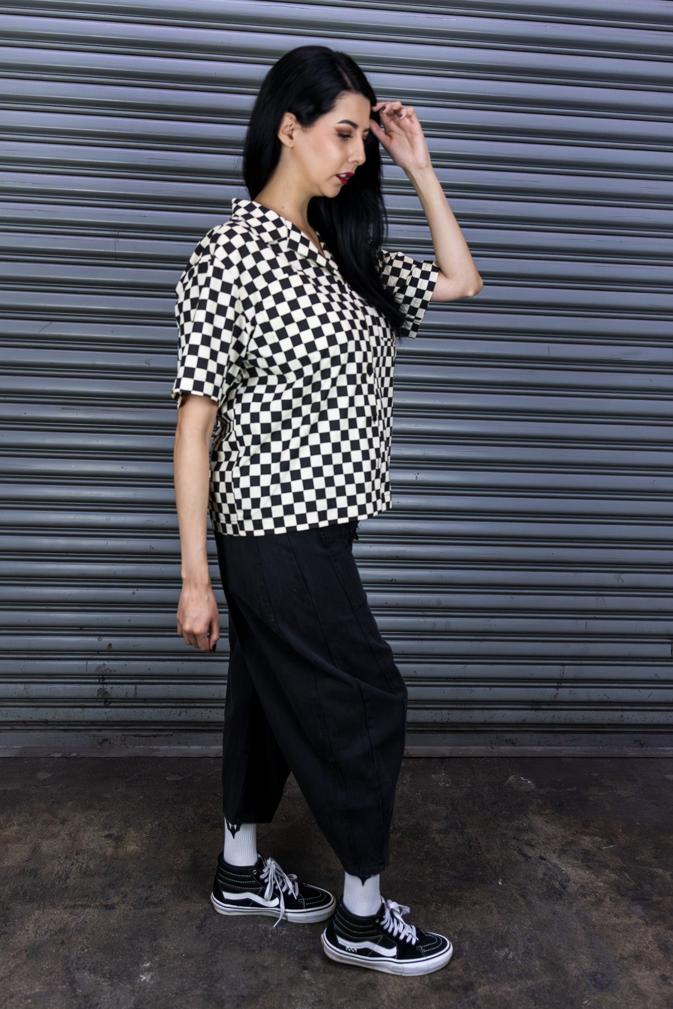 Person wearing a black and white checkered shirt with black pants in front of a metallic shutter.