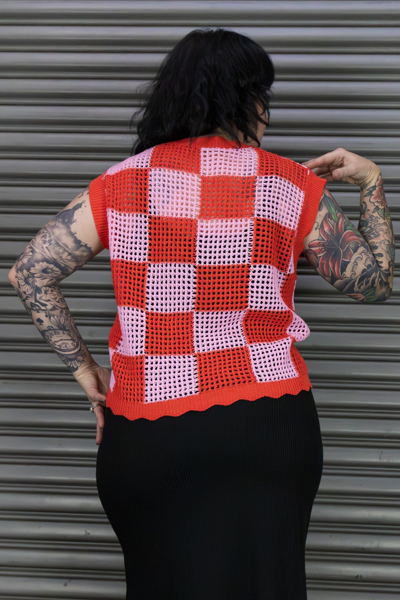 Person wearing a red and pink checkered crochet vest against a metal shutter background