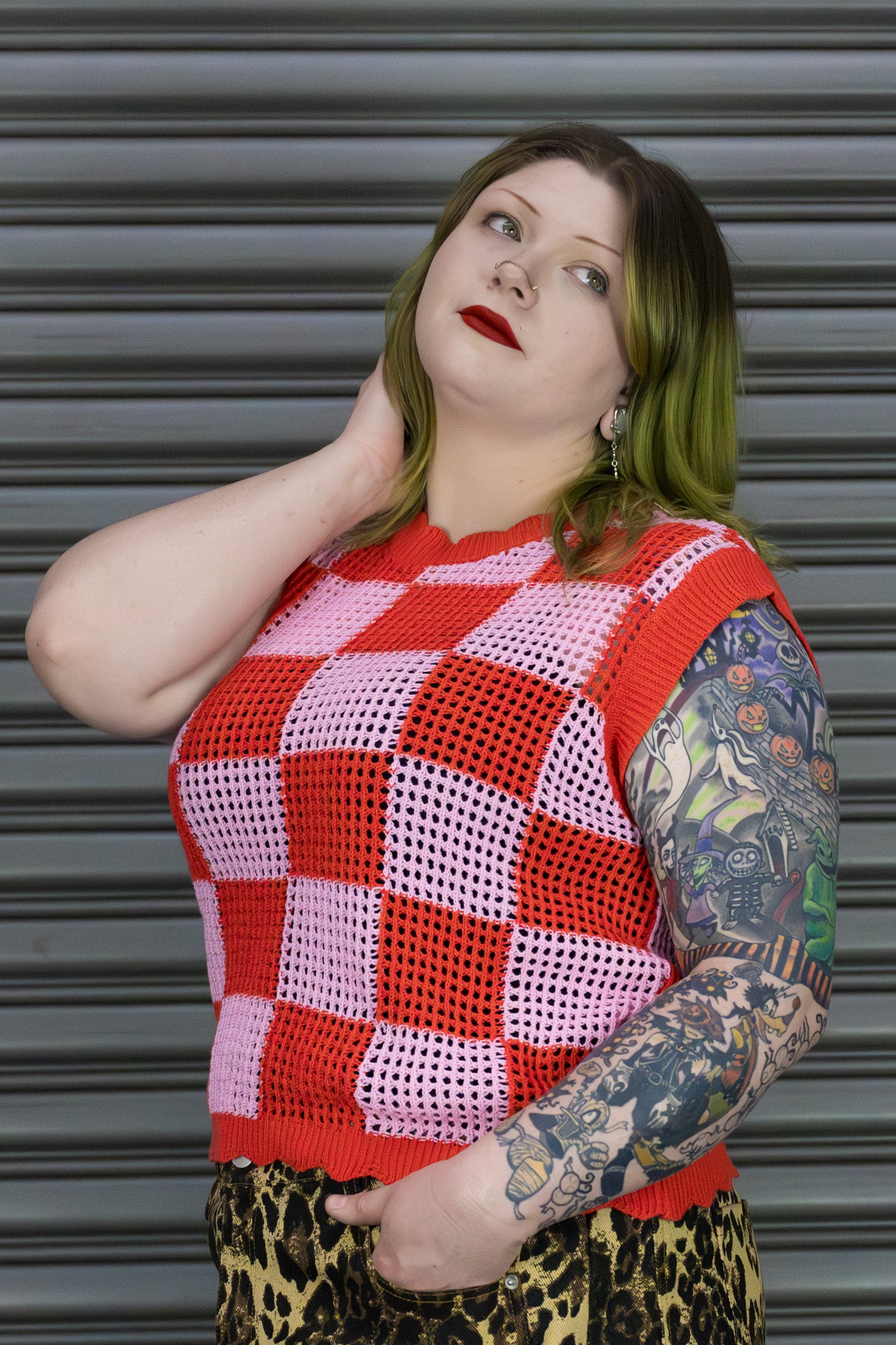 Person wearing a red and pink checkered sleeveless top with leopard print pants against a metallic shutter background.
