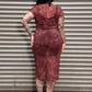 A woman with dark hair and visible tattoos stands with her back to the camera in front of a gray metal roll-up door, wearing a fitted short-sleeve midi dress in a rust red or terracotta color with a lighter floral or abstract pattern throughout.