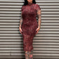 A woman with black hair and tattoos stands in front of a gray metal roll-up door, wearing a fitted short-sleeve high-neck dress with a red and black abstract pattern and black loafers.