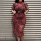 A woman with black hair and tattoos stands in front of a gray metal roll-up door, wearing a fitted burgundy leopard-print midi dress with a black belt, black shoulder harness straps, and black shoes.