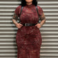 A woman with black hair and tattoos stands in front of a gray metal shutter, wearing a red and brown leopard-print dress with a black belt and suspenders.