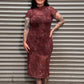 A woman with black hair and tattoos stands in front of a gray metal roll-up door, wearing a short-sleeve high-neck midi dress with a red and black abstract pattern and black shoes.