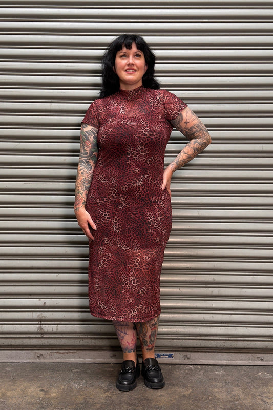 A woman with black hair and tattoos stands in front of a gray metal roll-up door, wearing a short-sleeve high-neck midi dress with a red and black abstract pattern and black shoes.