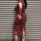 A woman with tattoos stands in front of a gray metal roll-up door, wearing a sleeveless, knee-length burgundy dress with a subtle gold pattern and black platform shoes.