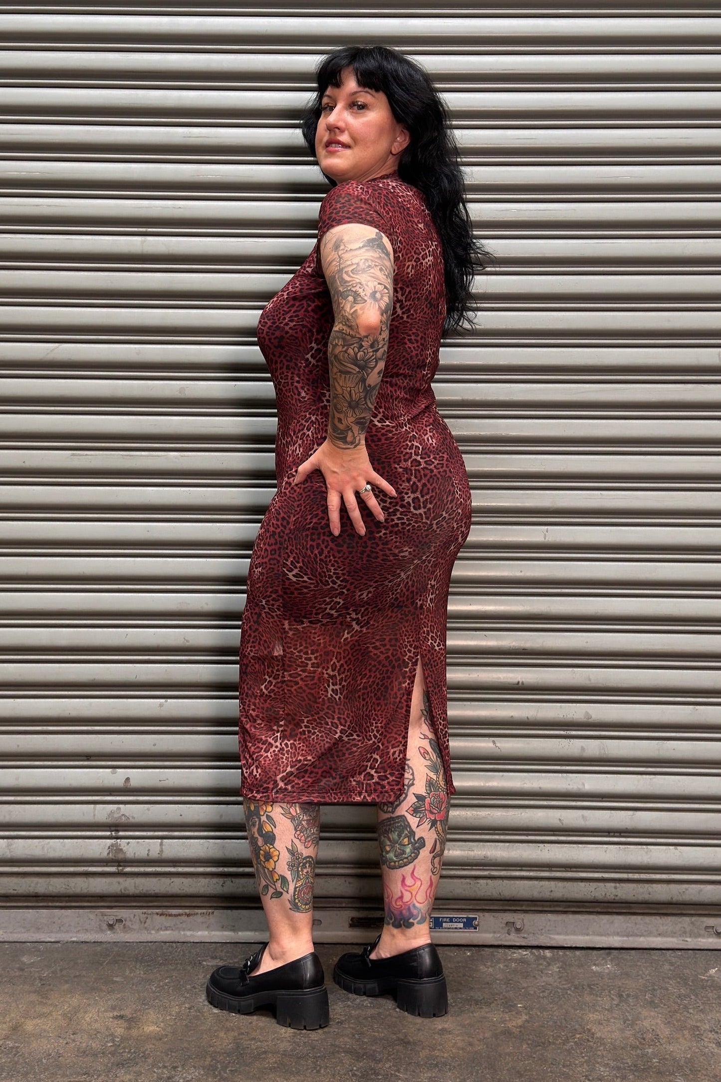 A woman with tattoos stands in front of a gray metal roll-up door, wearing a sleeveless, knee-length burgundy dress with a subtle gold pattern and black platform shoes.