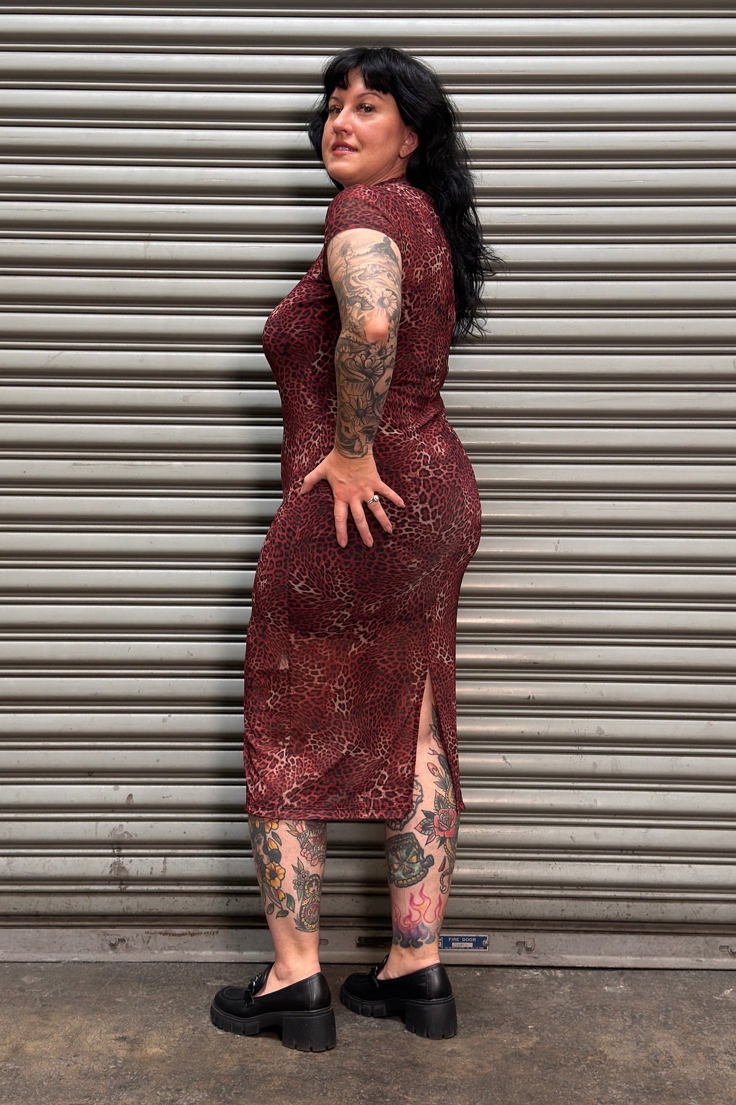 A woman with tattoos stands in front of a gray metal roll-up door, wearing a sleeveless, knee-length burgundy dress with a subtle gold pattern and black platform shoes.