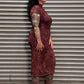 A woman with black hair and tattoos stands in profile against a gray metal roll-up door, wearing a fitted long-sleeve midi dress with a dark red and black abstract pattern and black loafers.