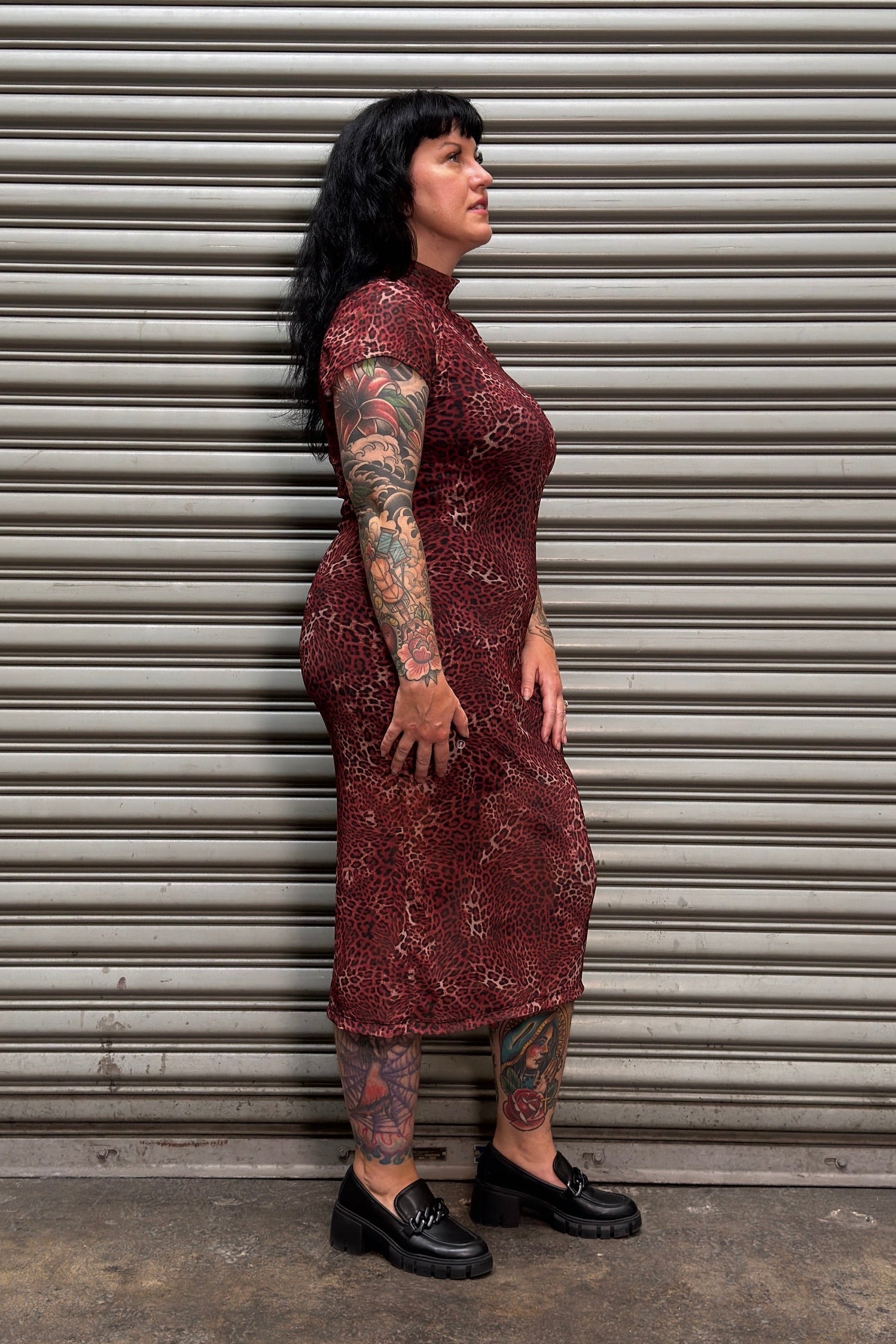 A woman with black hair and tattoos stands in profile against a gray metal roll-up door, wearing a fitted long-sleeve midi dress with a dark red and black abstract pattern and black loafers.