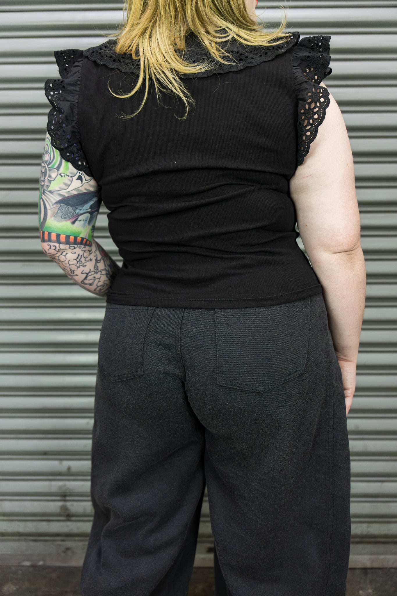 Person wearing a black sleeveless top with lace details and dark pants, standing against a metallic shutter.