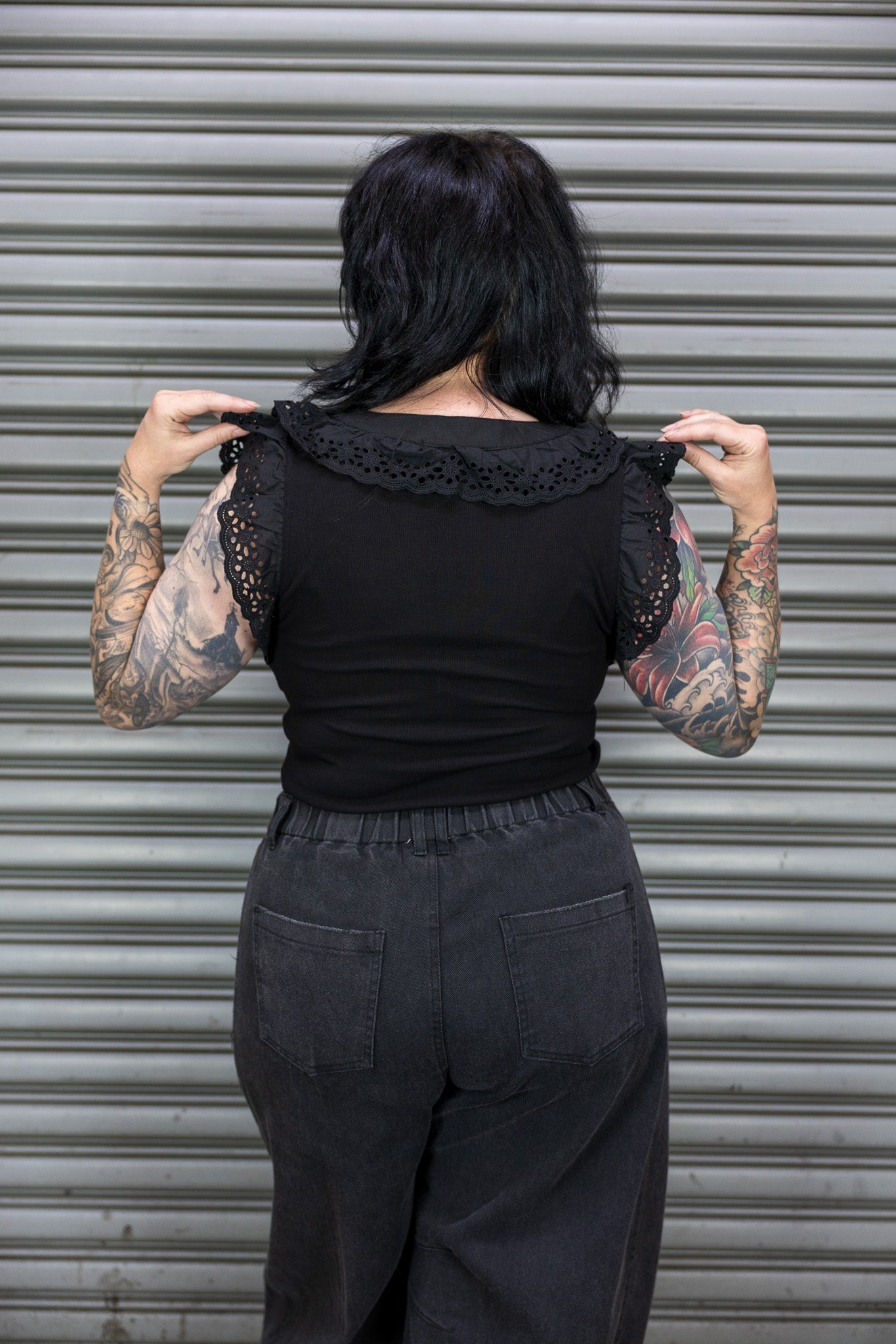 Person wearing a black top with lace details and dark pants in front of a metallic shutter.