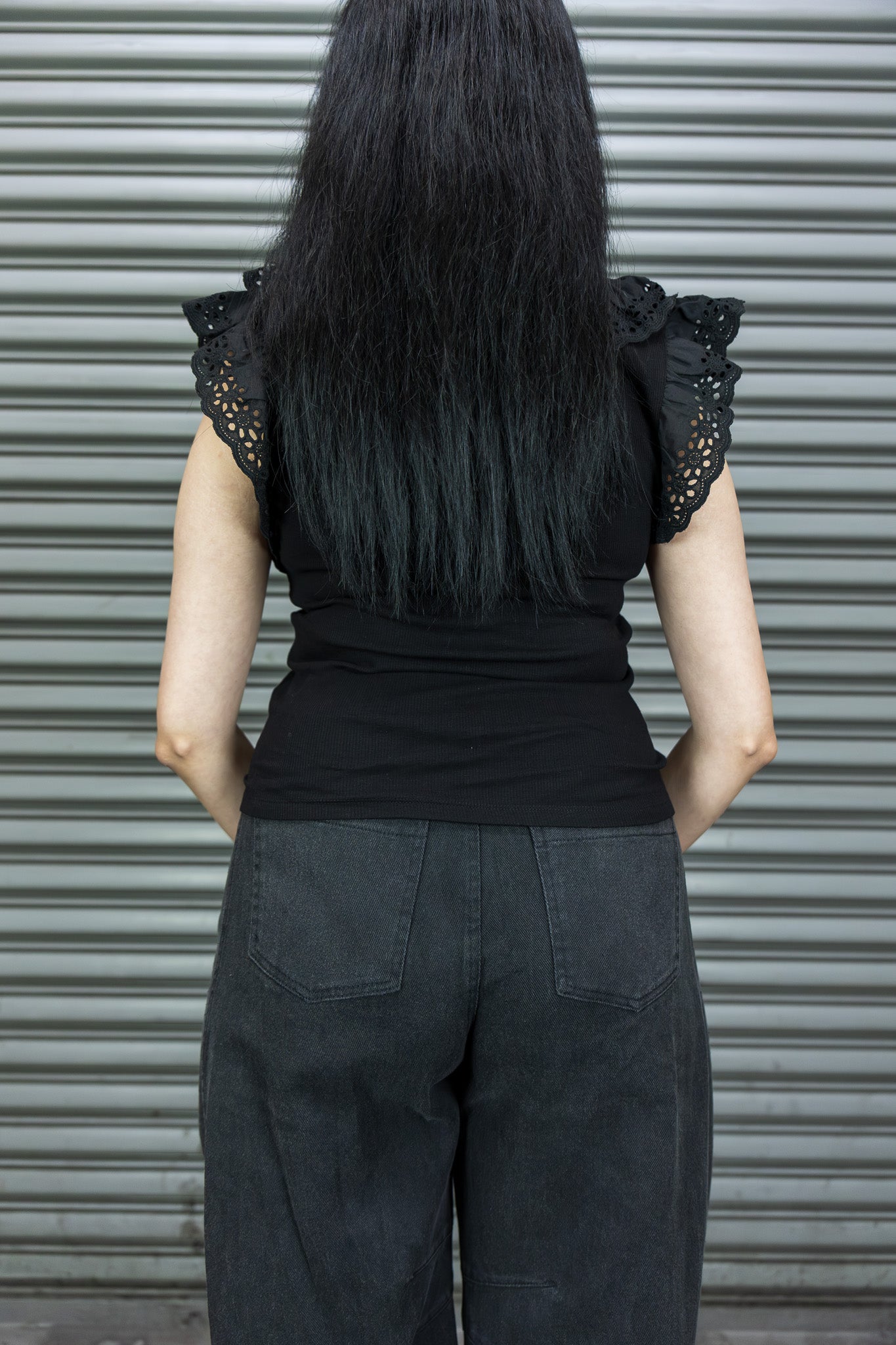 Person wearing a black top with ruffled sleeves and dark pants against a metallic shutter background