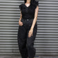 Woman wearing a black outfit with a ruffled top and high-waisted pants, standing in front of a metal shutter.