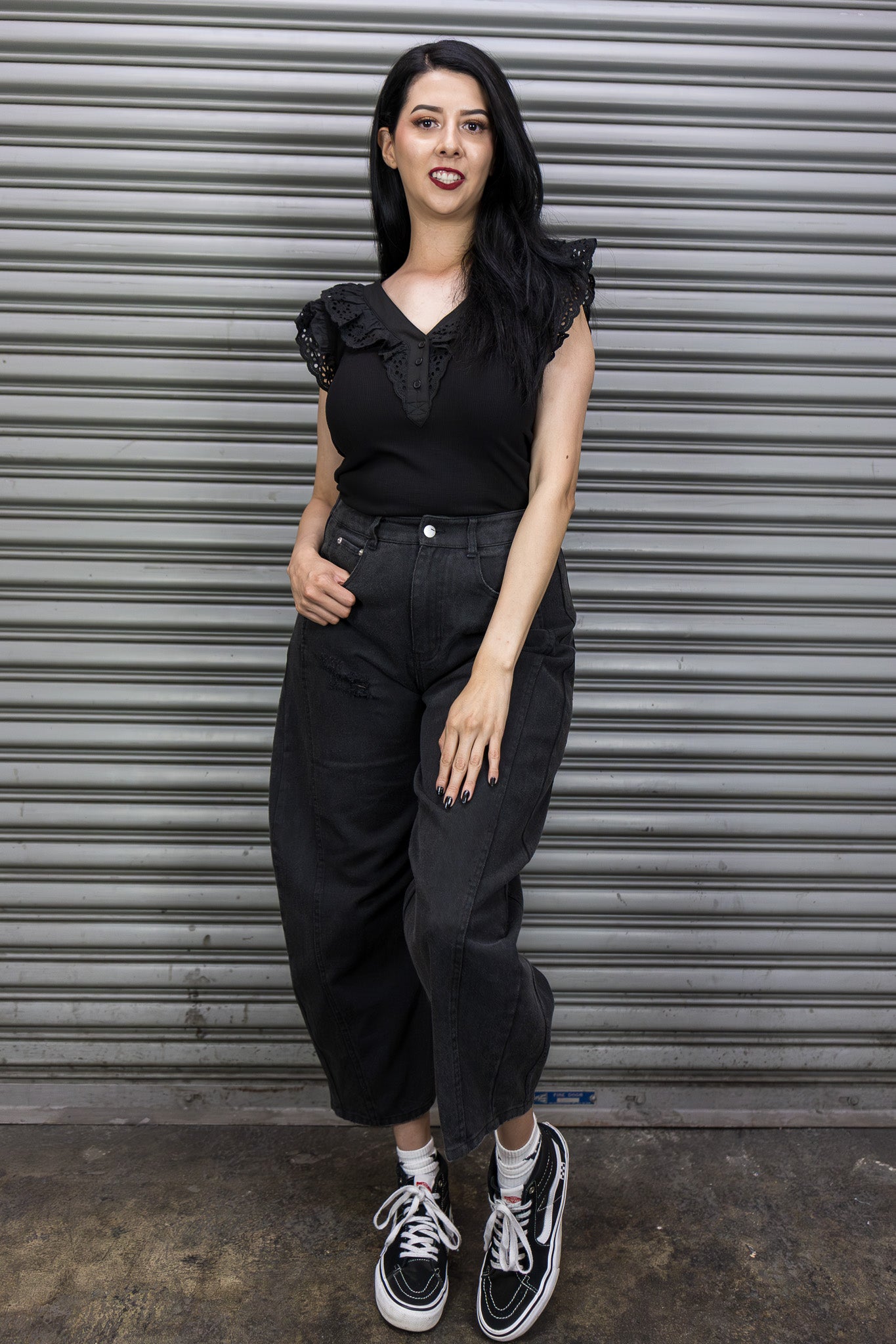 Woman wearing a black outfit with a ruffled top and high-waisted pants, standing in front of a metal shutter.