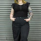 Person wearing a black sleeveless top with ruffled details and black pants against a metal shutter background.