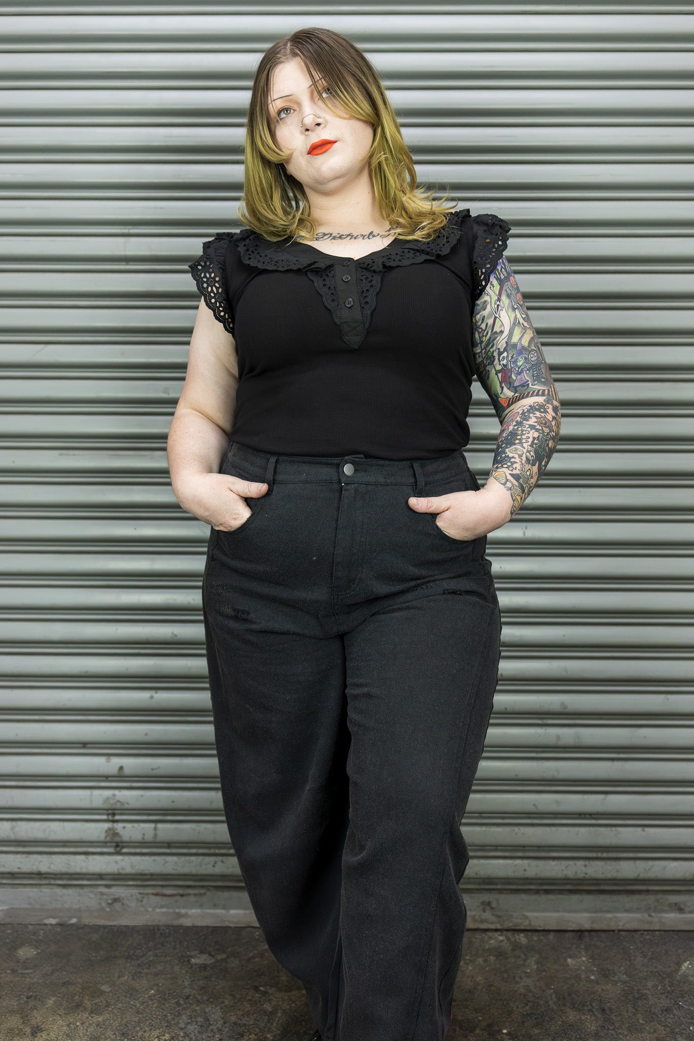 Person wearing a black sleeveless top with ruffled details and black pants against a metal shutter background.
