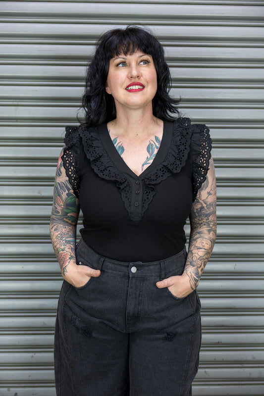 Woman wearing a black top with lace details and dark pants against a metal shutter background