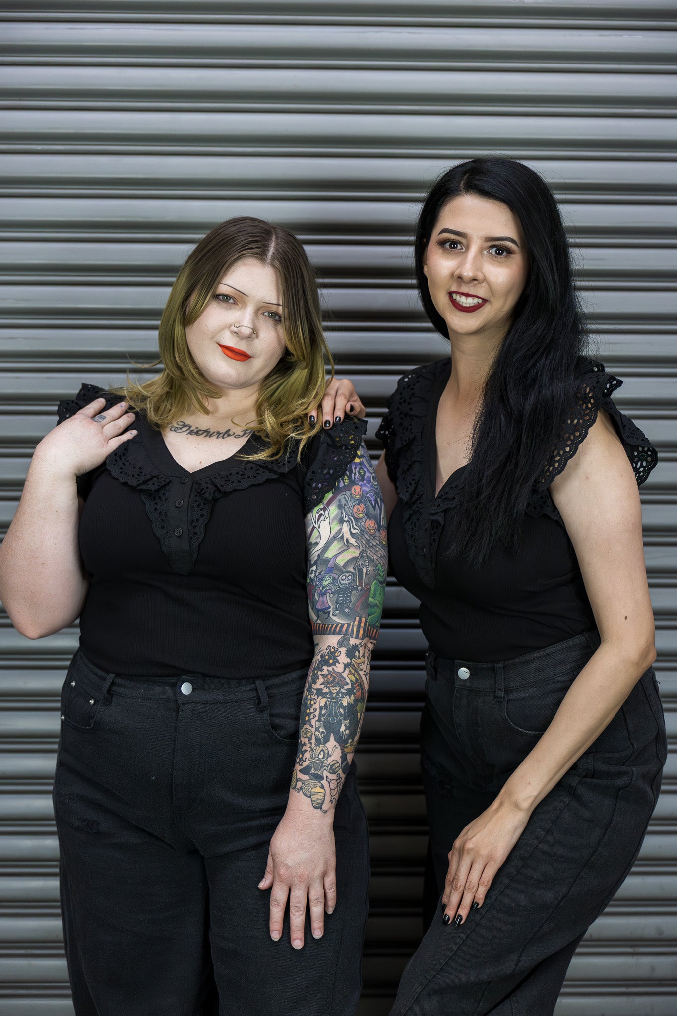 Two women in black outfits standing against a metallic shutter.