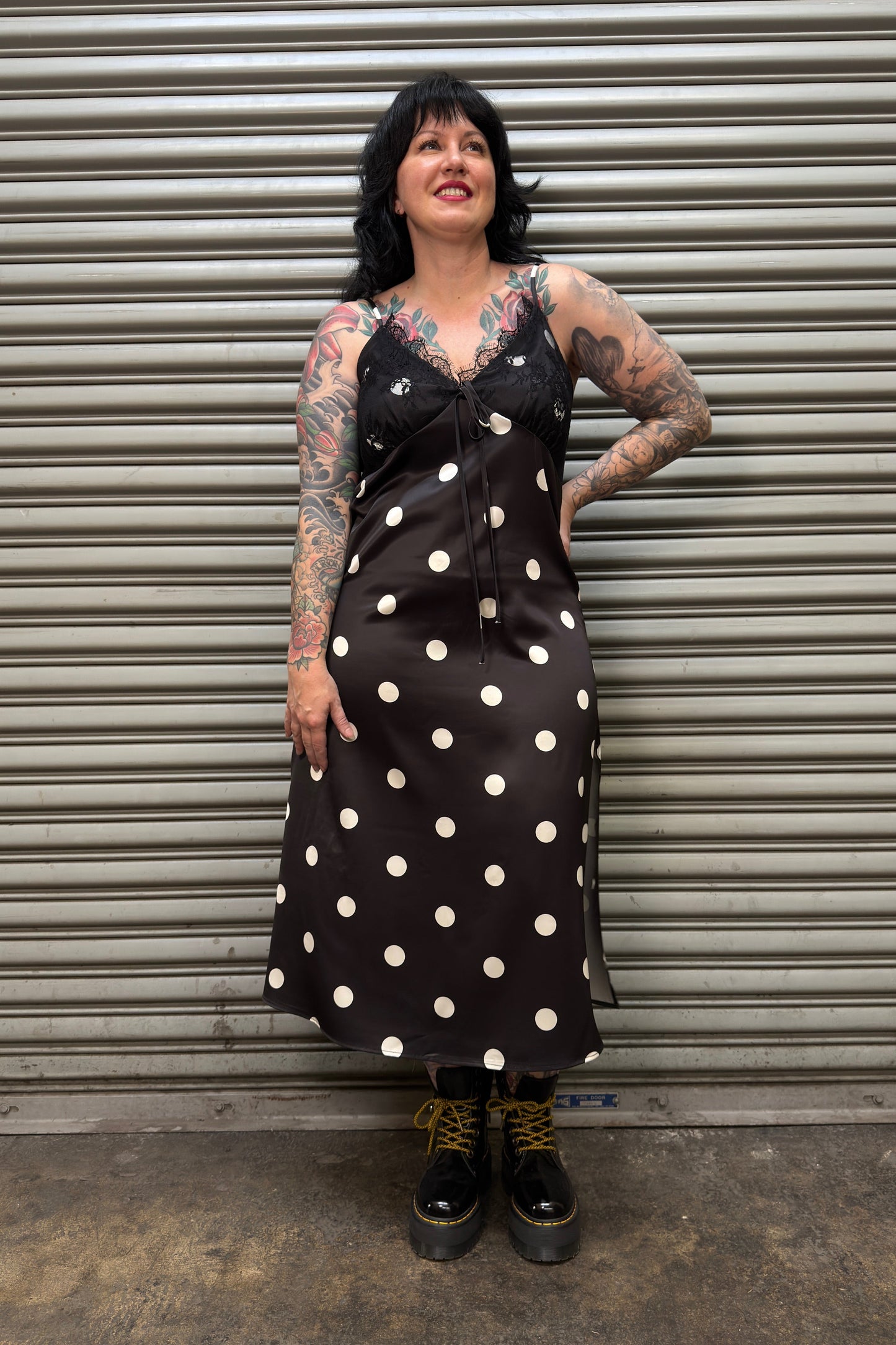 Woman wearing a black dress with white polka dots standing in front of a metal shutter.