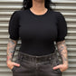 Woman wearing a black ribbed top and gray high-waisted pants standing in front of a metal shutter.