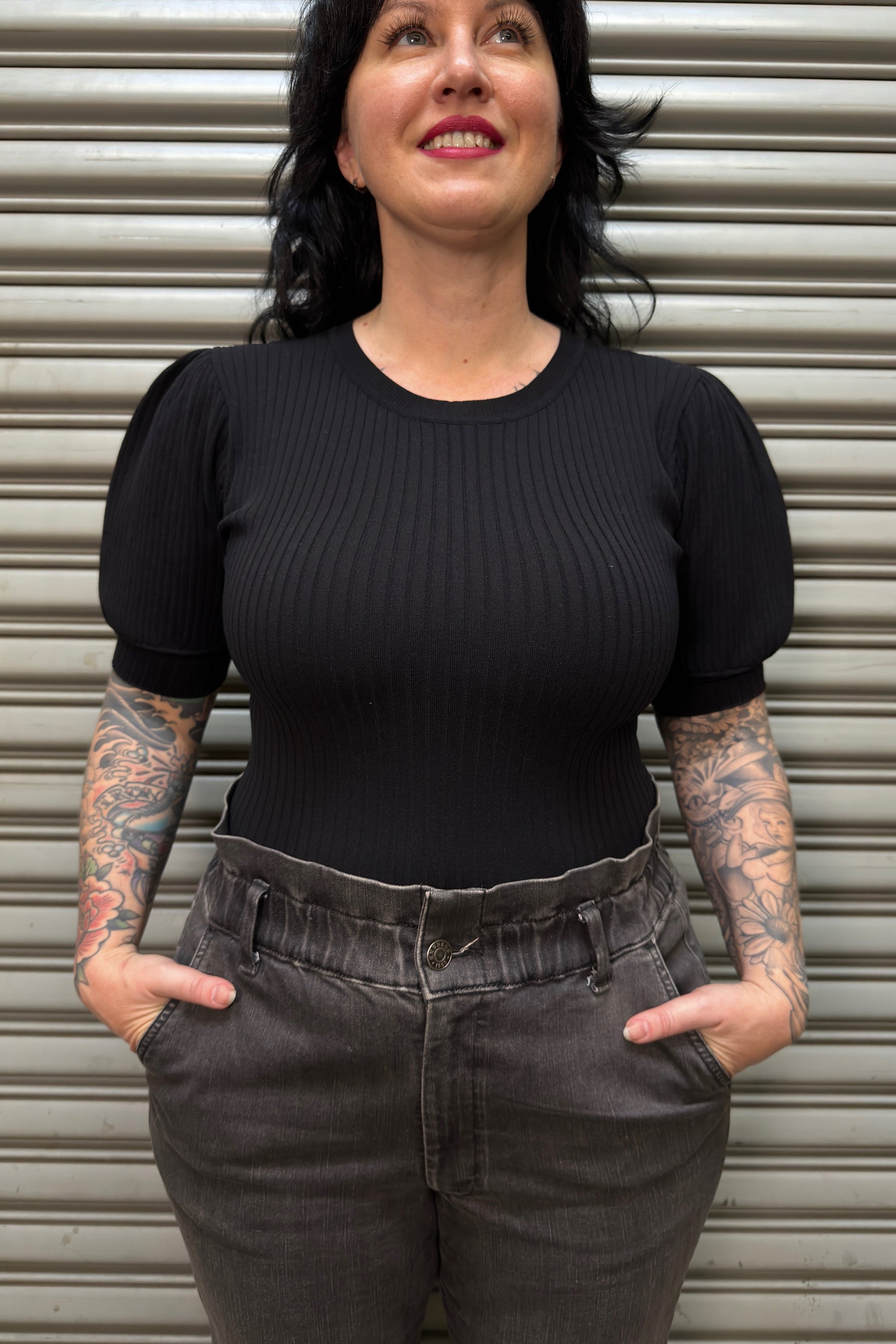 Woman wearing a black ribbed top and gray high-waisted pants standing in front of a metal shutter.