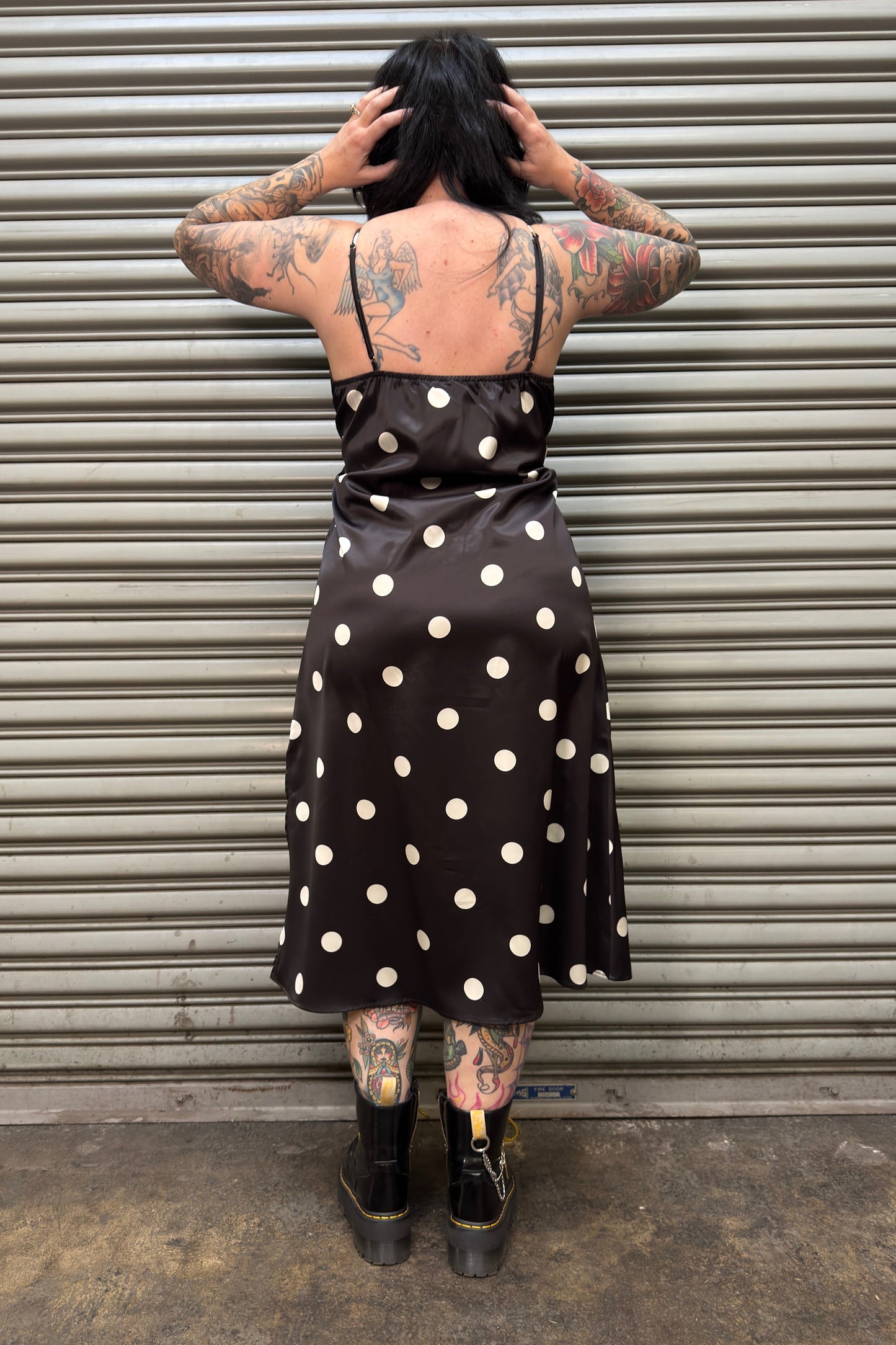 Person wearing a black polka dot dress standing in front of a corrugated metal wall.