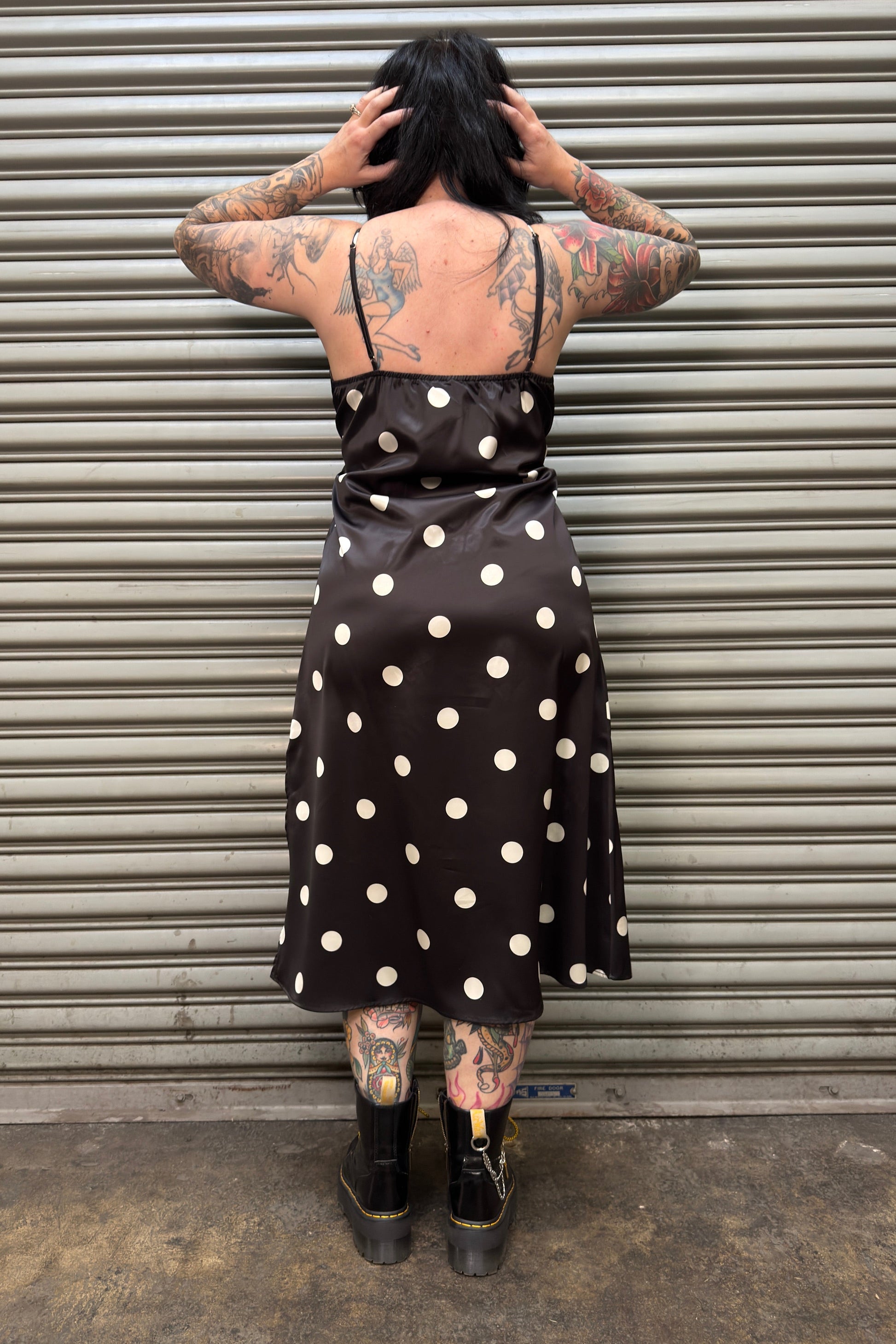 Person wearing a black polka dot dress standing in front of a corrugated metal wall.