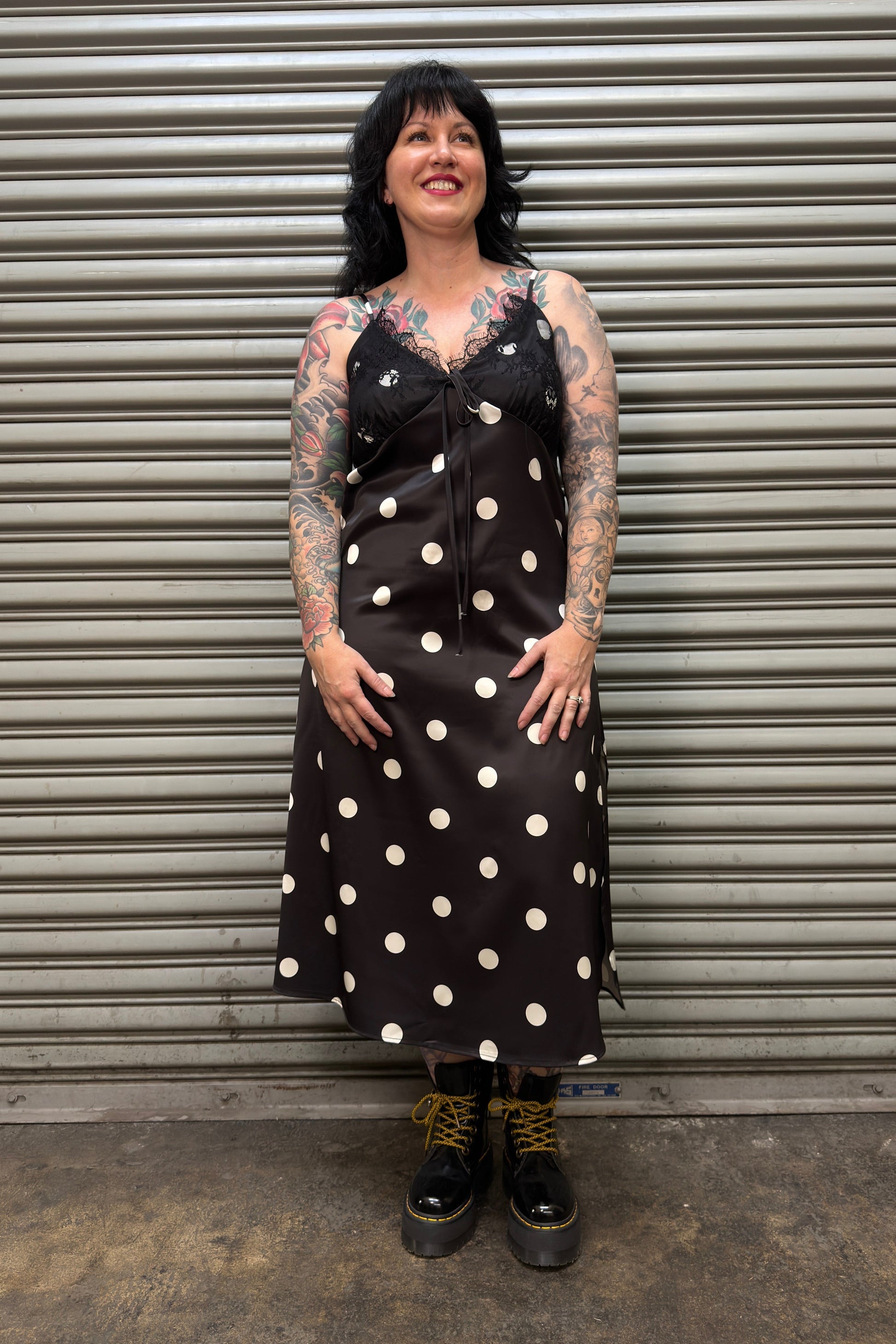 Woman wearing a black dress with white polka dots standing in front of a metal shutter.
