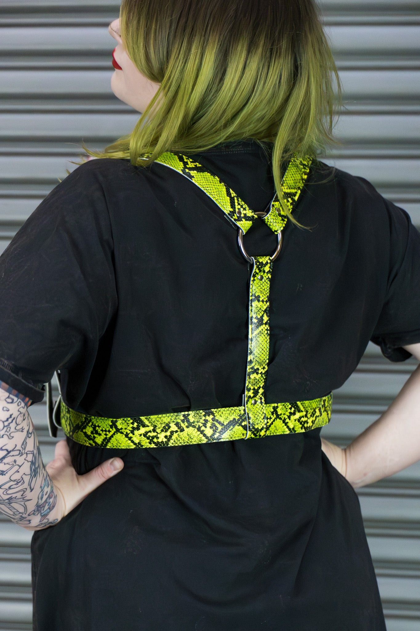 Person wearing a black outfit with a bright yellow and black patterned belt against a metallic background.