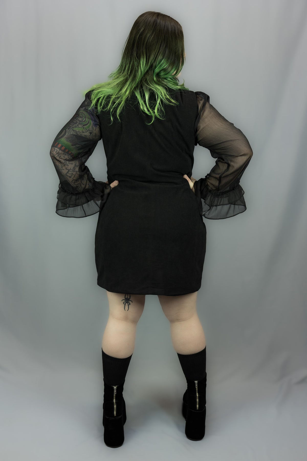 A model shown from the back wearing a black dress with sheer black bell sleeves, standing against a light gray studio backdrop.