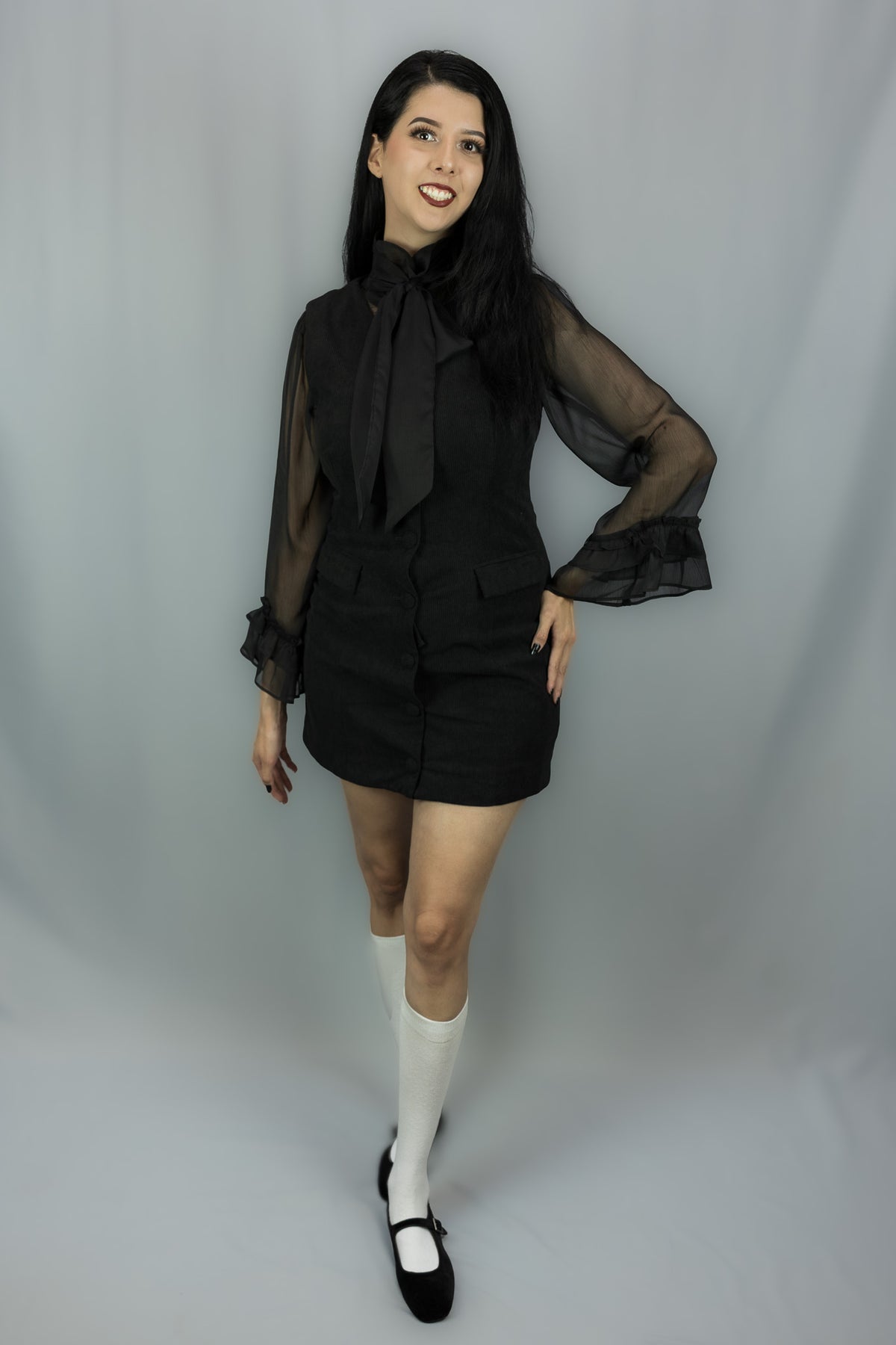 A woman modeling a black sheer long-sleeve blouse with a large bow at the collar and a matching black mini skirt, paired with white knee-high socks and black Mary Jane shoes, against a light gray studio backdrop.