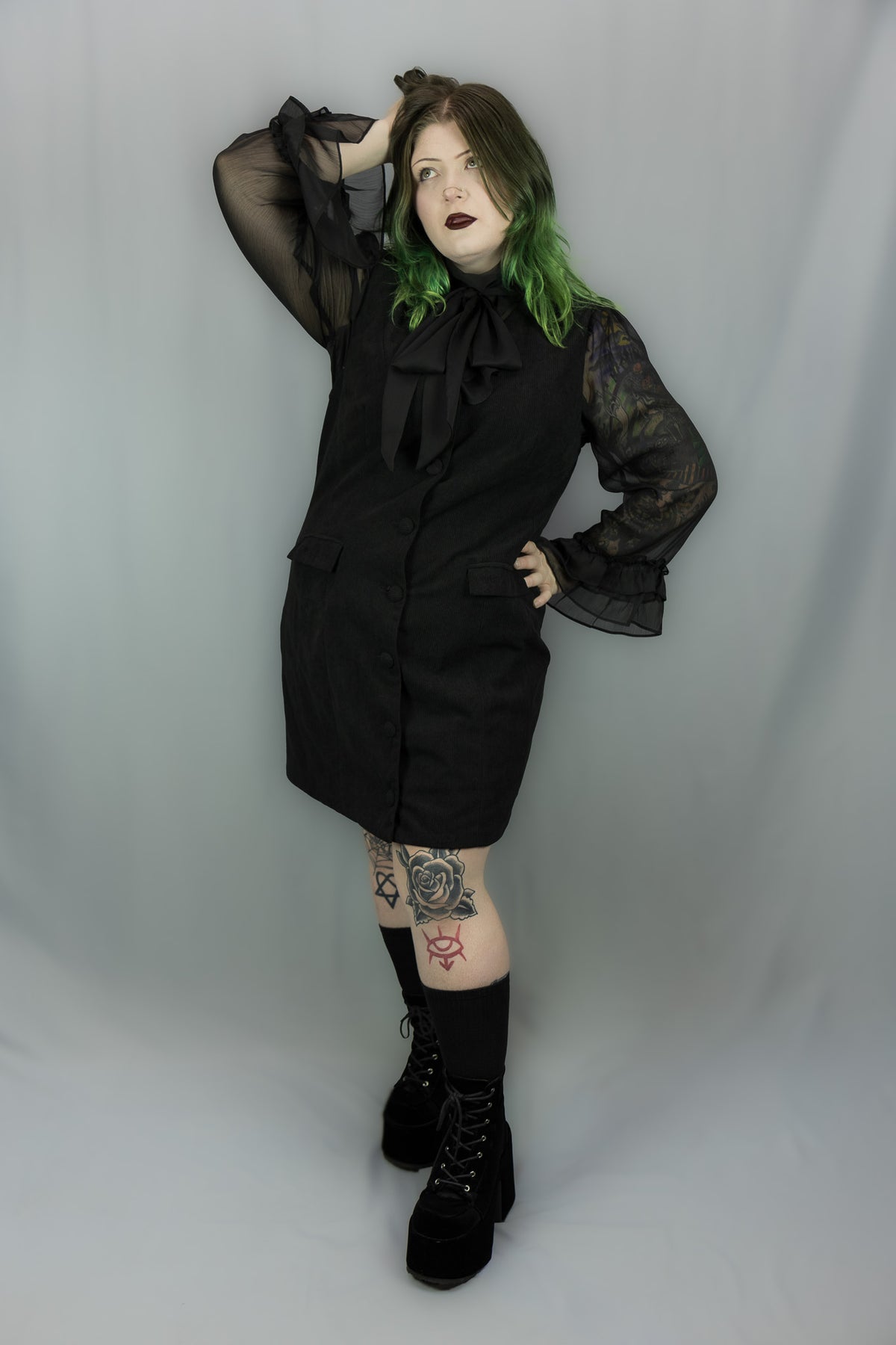 A model with green-tipped hair wearing a black gothic-style mini dress with sheer ruffled sleeves, a large black bow at the collar, black knee-high socks, and black platform boots, posing against a light gray studio backdrop.