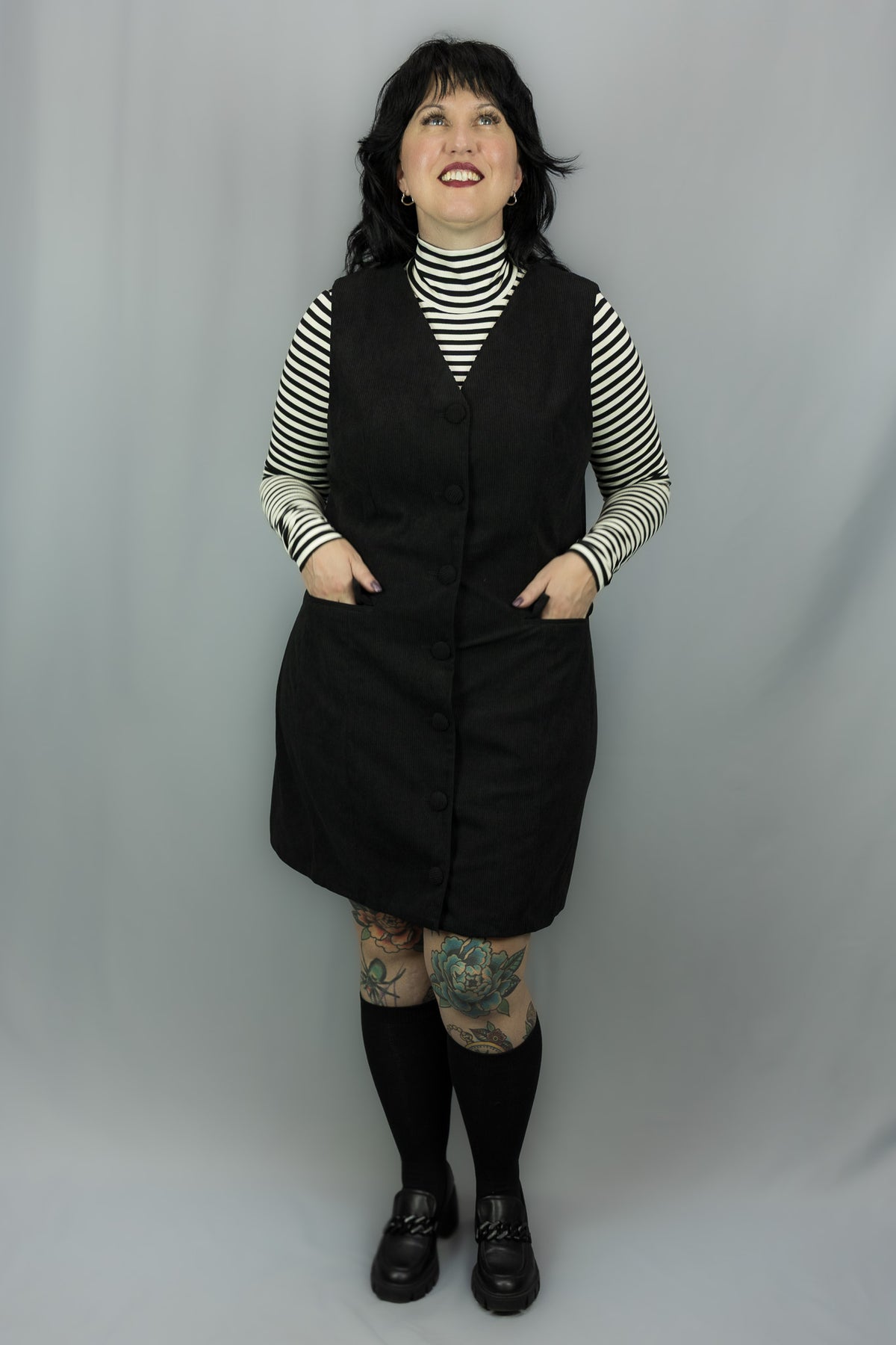 A woman modeling a black sleeveless dress over a black and white striped turtleneck, paired with black knee-high socks and black shoes, standing against a light gray studio backdrop.