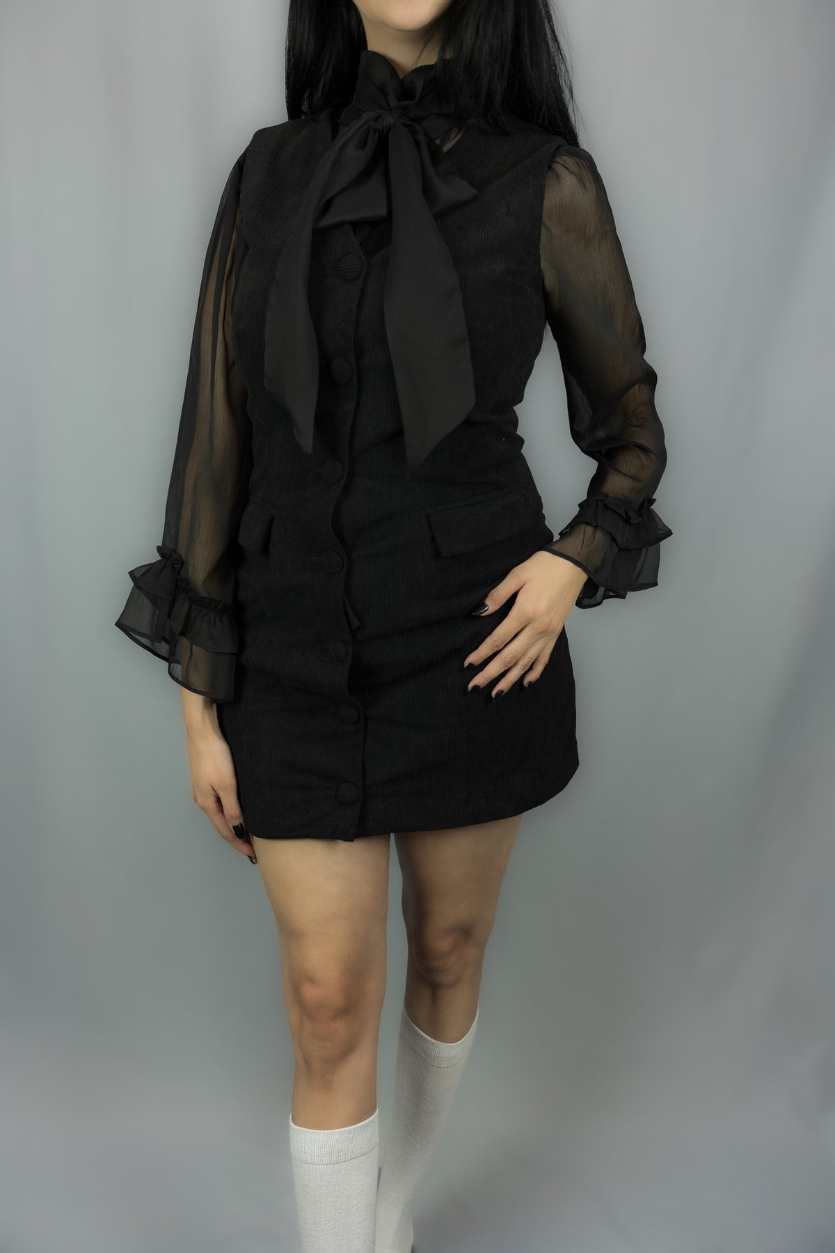 A model wearing a black gothic-style mini dress with sheer long sleeves, a large black bow at the collar, and white knee-high socks, photographed against a neutral gray studio background.