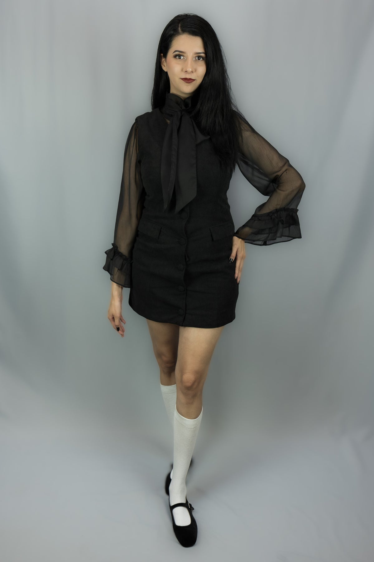 A woman modeling a black mini dress with sheer long sleeves, a high neck with a large bow, and a black waist tie, paired with white knee-high socks and black Mary Jane shoes, against a light gray studio backdrop.