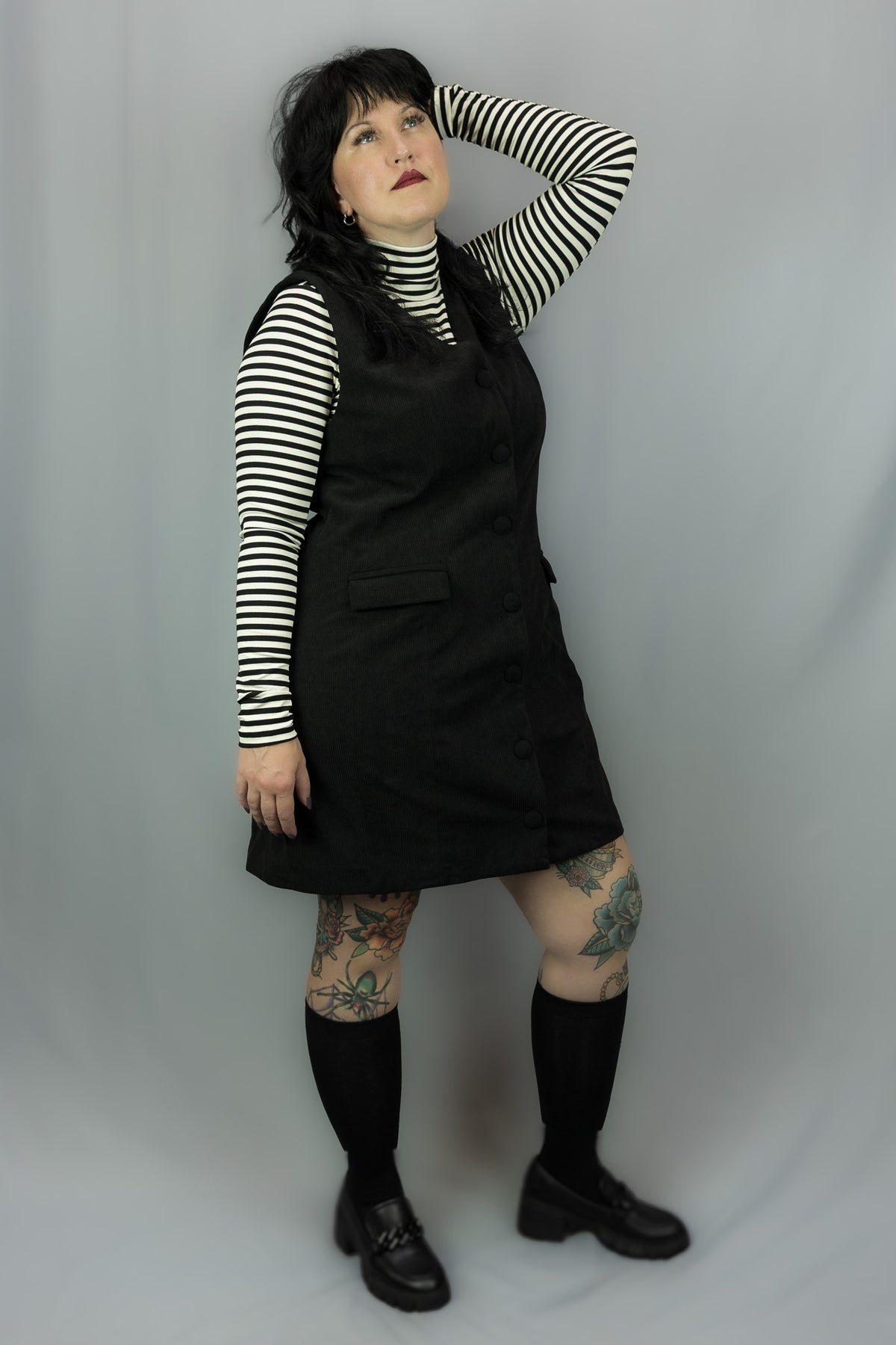 A person modeling a black dress over a black-and-white striped long-sleeve top, paired with black knee-high socks and black shoes, standing against a light gray studio backdrop.