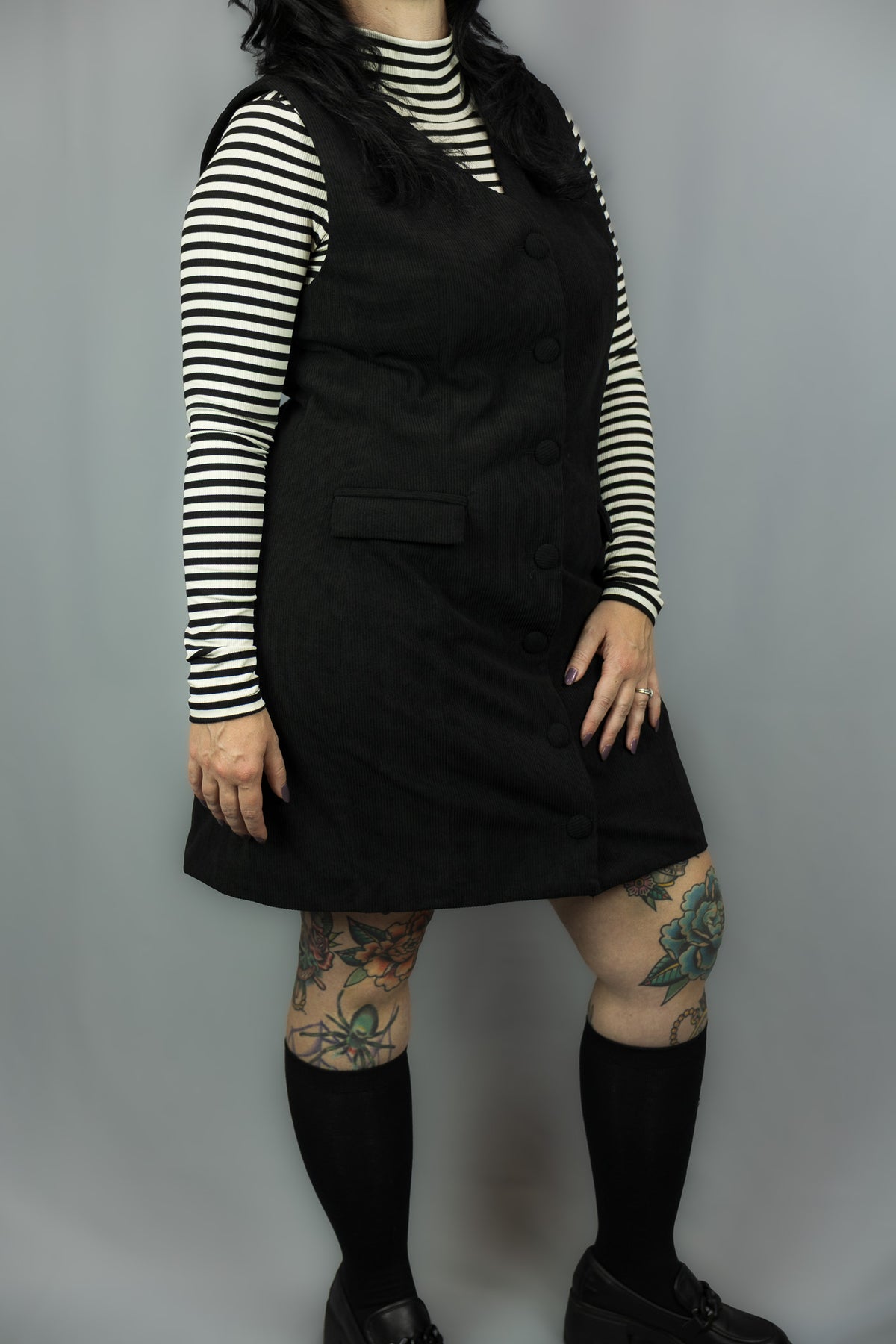 A person modeling a black sleeveless dress over a black-and-white striped long-sleeve top, paired with black knee-high socks and black shoes, against a plain gray studio background.